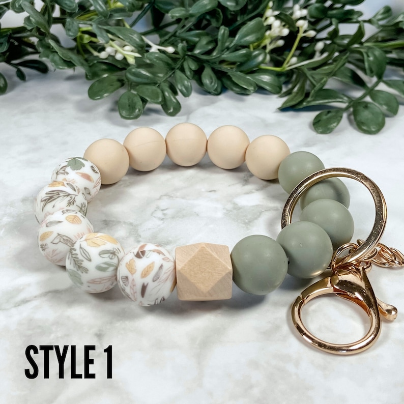 May include: A beige and green silicone beaded keyring with a gold metal key ring. The beads have a floral pattern and a wood bead.