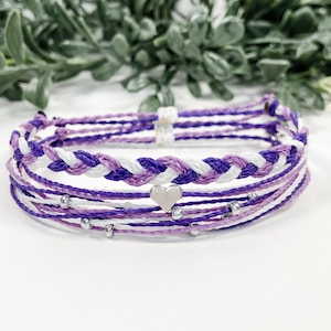 May include: A stackable bracelet set featuring shades of purple and white. The bracelets have a braided design and a small silver heart charm. The bracelets are made of string and have small silver beads.