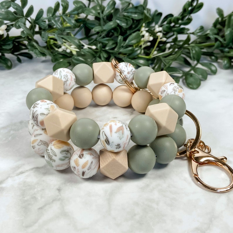 May include: A green and beige silicone beaded keyring with a floral pattern. The keyring has a gold metal clasp.
