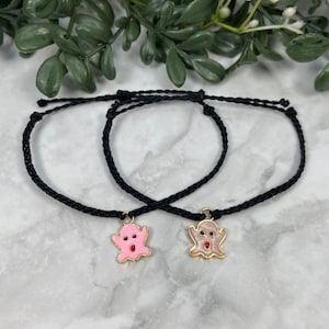 May include: Two black friendship bracelets with ghost charms. One charm is pink with a red mouth and the other is beige with a red mouth.