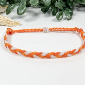May include: A white and orange braided friendship bracelet. The bracelet is made of thin string and has a simple knot closure.