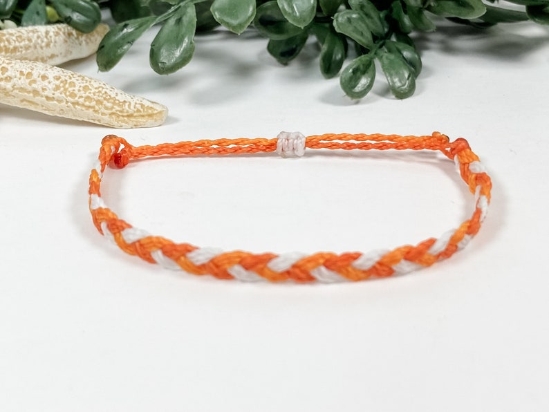 May include: A white and orange braided friendship bracelet. The bracelet is made of thin string and has a simple knot closure.