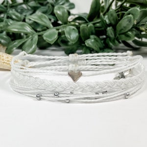 May include: White braided friendship bracelets with silver beads and a silver heart charm. The bracelet has the word "HOPE" written on it.