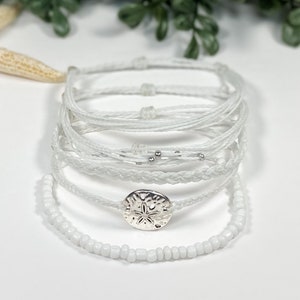 May include: A set of white bracelets with a silver starfish charm. The bracelets are made of different materials, including string, beads, and braided cord.