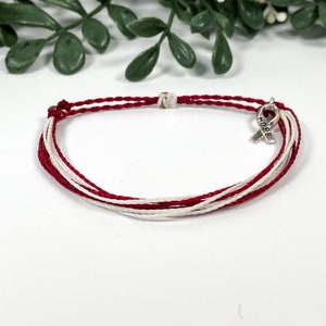 May include: Red and white braided bracelet with a silver charm in the shape of a ribbon with the word "HOPE" on it.