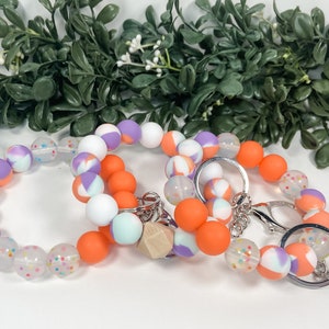 May include: A set of three silicone beaded bracelets in orange, white, purple, and light blue with a silver key chain. The bracelets have a speckled pattern and the key chain has a wooden bead.