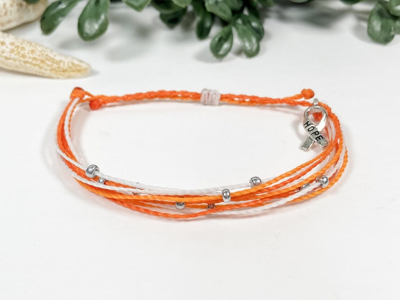 May include: Orange and white friendship bracelet with silver beads and a silver charm that says "HOPE" in all caps.