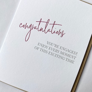 May include: A white card with the word "congratulations" written in burgundy script. The text "You're engaged! Enjoy every moment of this exciting time" is printed in gray below the word "congratulations".