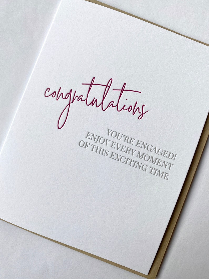May include: A white card with the word "congratulations" written in burgundy script. The text "You're engaged! Enjoy every moment of this exciting time" is printed in gray below the word "congratulations".