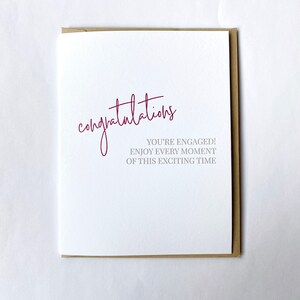 May include: A white card with the word "congratulations" written in pink cursive. The text "You're engaged! Enjoy every moment of this exciting time" is written in gray below the word "congratulations".