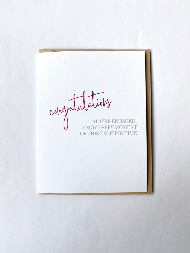 May include: A white card with the word "congratulations" written in pink cursive. The text "You're engaged! Enjoy every moment of this exciting time" is written in gray below the word "congratulations".