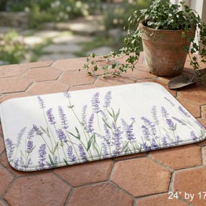 May include: A rectangular bath mat with a lavender floral design. The mat is white with purple lavender flowers and green stems. The mat measures 24 inches by 17 inches.