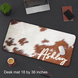 May include: A brown and white cowhide desk mat with the name "Ashley" written in white. The mat is 18 by 36 inches.