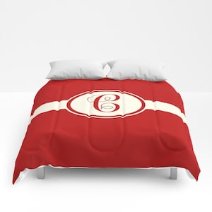 May include: A red comforter featuring a horizontal cream-coloured band across the centre. In the middle of the band is a cream-coloured oval frame with a red border, containing a large, stylised red letter C in a cursive script font.