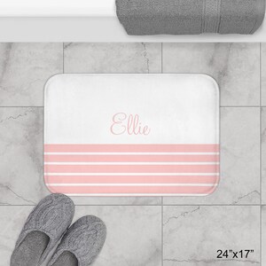 May include: White bath mat with pink stripes and the name "Ellie" printed in pink script. The mat measures 24 inches by 17 inches.