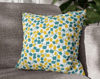 May include: Square throw pillow with a white background and a repeating pattern of teal and yellow leaf shapes. The pillow is resting on a gray sofa. The leaves are connected by thin, curving lines.