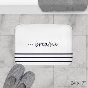 May include: White rectangular bath mat with the text "... breathe" in a cursive font. The mat has three navy blue stripes at the bottom and measures 61cm x 43cm. A pair of gray knitted slippers are in the foreground.