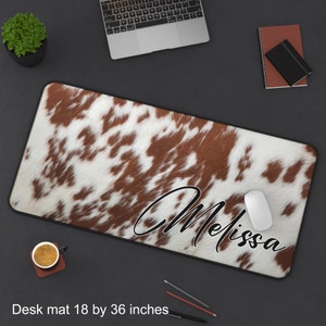 May include: A brown and white cowhide desk mat with a black border. The mat is 18 by 36 inches and has the name "Melissa" written in a cursive font on it.