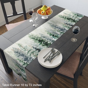 May include: A rectangular table runner with a watercolour design of evergreen trees and falling snow. The runner is 40.6 x 182.9 cm. The runner is placed on a dark wooden table, set with plates, forks, wine glasses, and a bowl of apples.