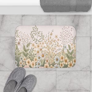 May include: A rectangular bath mat featuring a soft pink background with a dense, repeating pattern of delicate wildflowers and leafy stems in shades of sage green, cream, and muted yellow, arranged in a dense, vertical botanical composition.