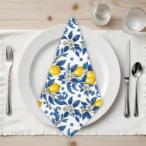 May include: A white cloth napkin folded into a triangle and placed on a white dinner plate. The napkin features a repeating pattern of bright yellow lemons, white citrus blossoms, and deep blue leaves on a crisp white background.