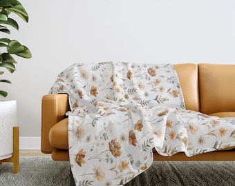 May include: A soft, white throw blanket with a floral pattern in shades of brown and beige. The blanket is draped over a brown leather couch.