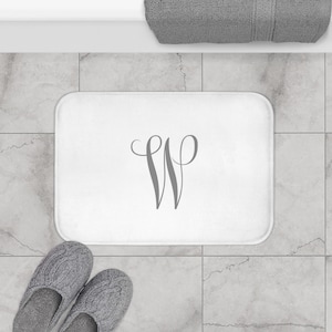 May include: A white rectangular bath mat with a gray cursive letter "W" in the center. The mat is on a tiled floor, with a gray towel and a pair of gray slippers visible.