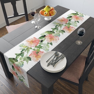 May include: White table runner with a watercolor floral design featuring large pink and peach flowers, green leaves, and blue accents. The runner is on a dark wood table, with plates, silverware, and wine glasses.