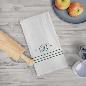 May include: White kitchen towel with a green embroidered letter "B" and two green stripes. The towel is on a wooden surface with a rolling pin, apples, and a mug.