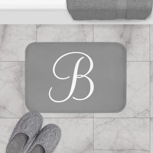 May include: Grey bath mat with a white cursive letter B in the centre.