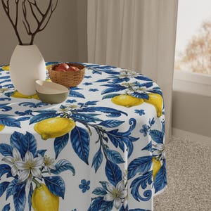 May include: A round tablecloth featuring a repeating pattern of bright yellow lemons, white citrus blossoms, and deep blue leaves on a white background. The fabric is draped over a circular table, with a white vase and a small woven bowl on top.
