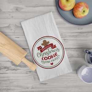 May include: White kitchen towel with a circular design that reads "OFFICIAL Christmas COOKIE TESTING FACILITY" in green and red. A gingerbread man and a red banner are also featured in the design. A wooden rolling pin is on the left.