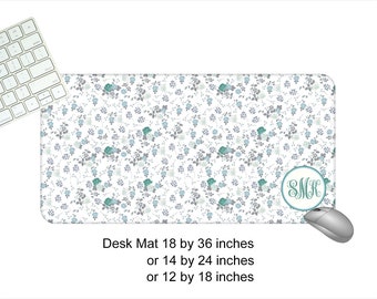 May include: A white desk mat with a floral pattern in shades of blue and gray. The mat has a monogram in a circle in the lower right corner that says "SMK". The mat is available in three sizes: 18 by 36 inches, 14 by 24 inches, and 12 by 18 inches.