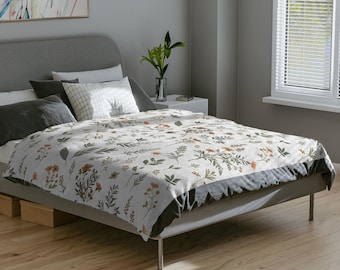 May include: A white floral blanket, measuring 80" x 60", covers a bed. The blanket features a pattern of green and orange botanical elements. The bed has a gray headboard and a dark gray bed skirt. The room has a neutral color palette.