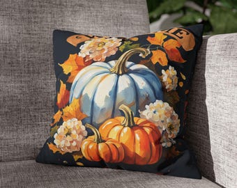 May include: A square throw pillow featuring a dark background with a large light blue pumpkin and two smaller orange pumpkins. The design includes clusters of white and cream flowers and scattered orange autumn leaves. A partial banner is visible at the top.