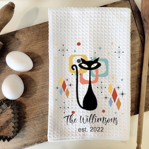 May include: White kitchen towel with a black cat illustration in a retro style. The cat is surrounded by colorful geometric shapes and stars. The text "The Williamsons est. 2022" is printed below the cat.