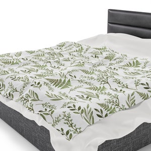 May include: A white throw blanket with a green leafy pattern. The blanket is draped over a grey upholstered bed.