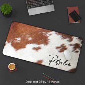 May include: A brown and white cowhide desk mat with the name "Rosalie" written in black cursive. The mat measures 36 by 18 inches.