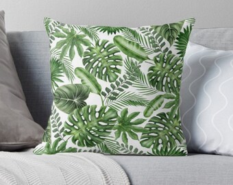 May include: A square throw pillow featuring a vibrant green tropical leaf pattern on a white background. The design includes various leaf shapes, such as Monstera, palm fronds, and other tropical foliage. The pillow is placed on a gray sofa.