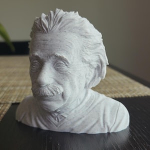 May include: A light gray 3D printed bust of Albert Einstein featuring his signature wild hair and mustache. The sculpture captures the facial features and texture of the physicist in a monochromatic matte finish, presented as a head and shoulder portrait.