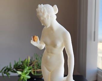May include: A white sculpture of a nude woman holding a small, round, golden-brown object in her right hand. She is also holding a draped cloth. The sculpture is on a black surface.
