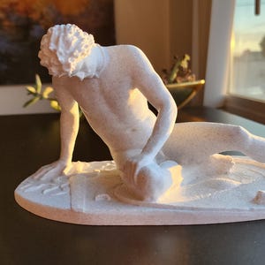 May include: A white sculpture of a reclining figure, possibly a classical statue. The figure is lying on a textured base with a curved design. The sculpture is lit by natural light, highlighting its form and details.