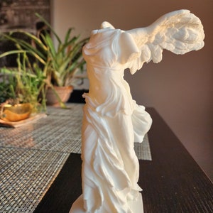 May include: A white sculpture of the Winged Victory of Samothrace, a classical Greek statue. The statue depicts a draped, winged figure with detailed feathering on the wings. The sculpture is mounted on a rectangular base and stands on a dark surface.