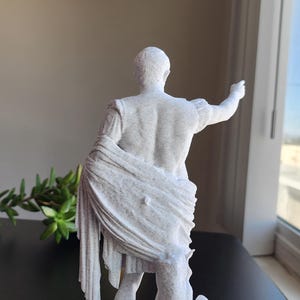 May include: A white, speckled, textured statue of a figure in classical attire, viewed from behind. The figure has one arm extended outward and is draped in a long, flowing garment that falls to the base, standing on a square pedestal.