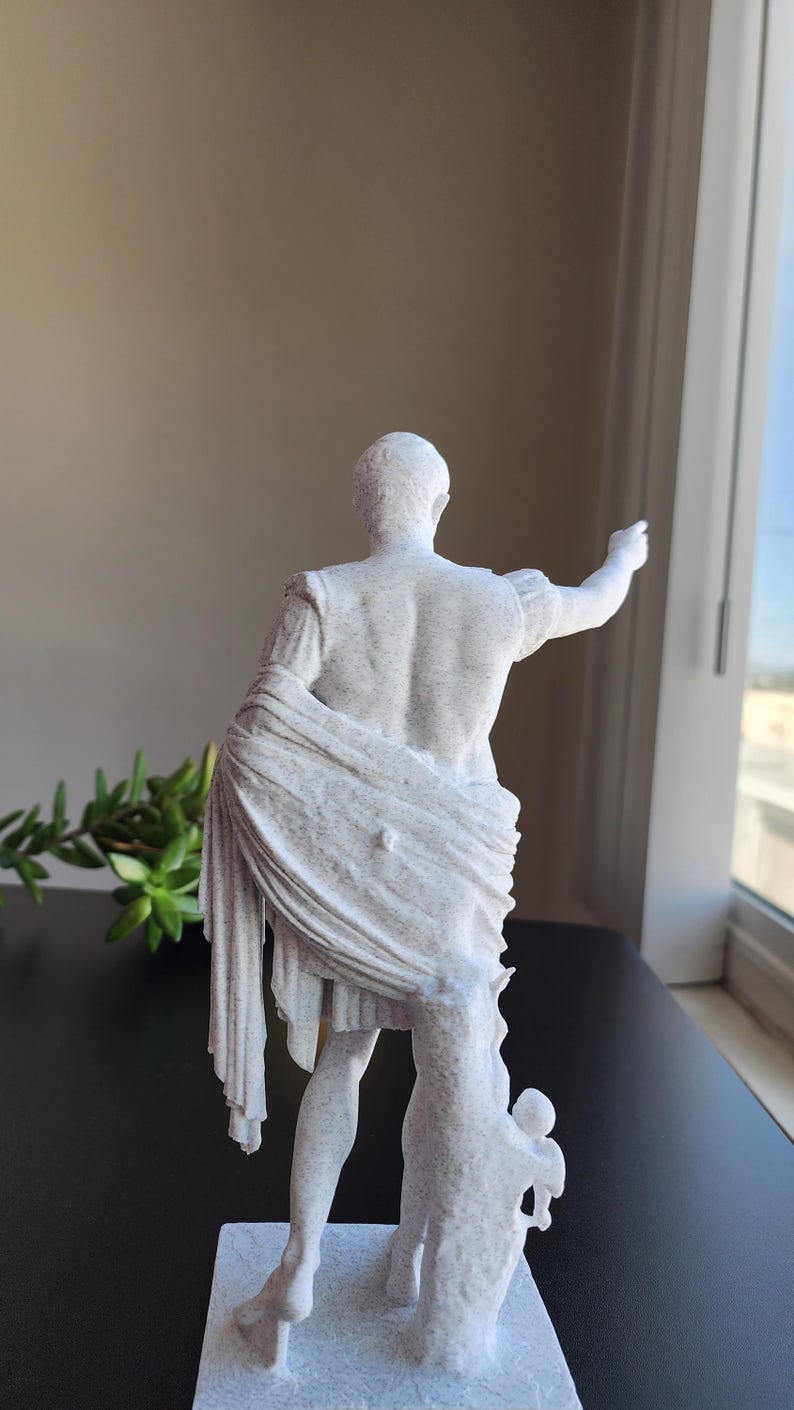May include: A white, speckled, textured statue of a figure in classical attire, viewed from behind. The figure has one arm extended outward and is draped in a long, flowing garment that falls to the base, standing on a square pedestal.