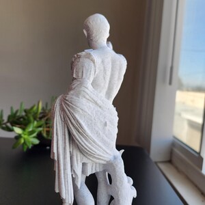 May include: A white, speckled, textured statue of a figure standing on a rectangular base. The figure is depicted from the back, wearing a draped garment over one shoulder that falls to the legs, with a tree-like support structure at the side.