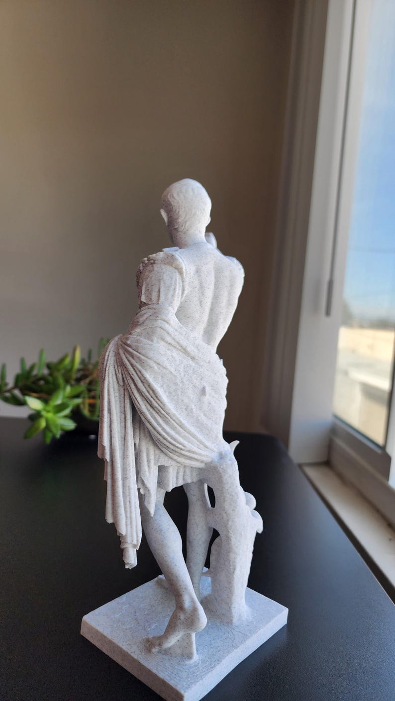 May include: A white, speckled, textured statue of a figure standing on a rectangular base. The figure is depicted from the back, wearing a draped garment over one shoulder that falls to the legs, with a tree-like support structure at the side.