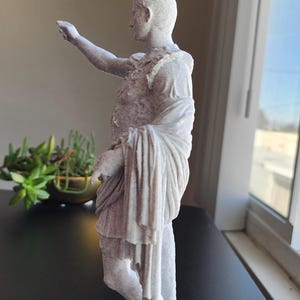 May include: A light-colored, speckled statue of a figure in Roman-style armor and a draped toga. The figure stands on a rectangular base with one arm extended forward and the other arm bent at the side, featuring detailed fabric folds and textures.