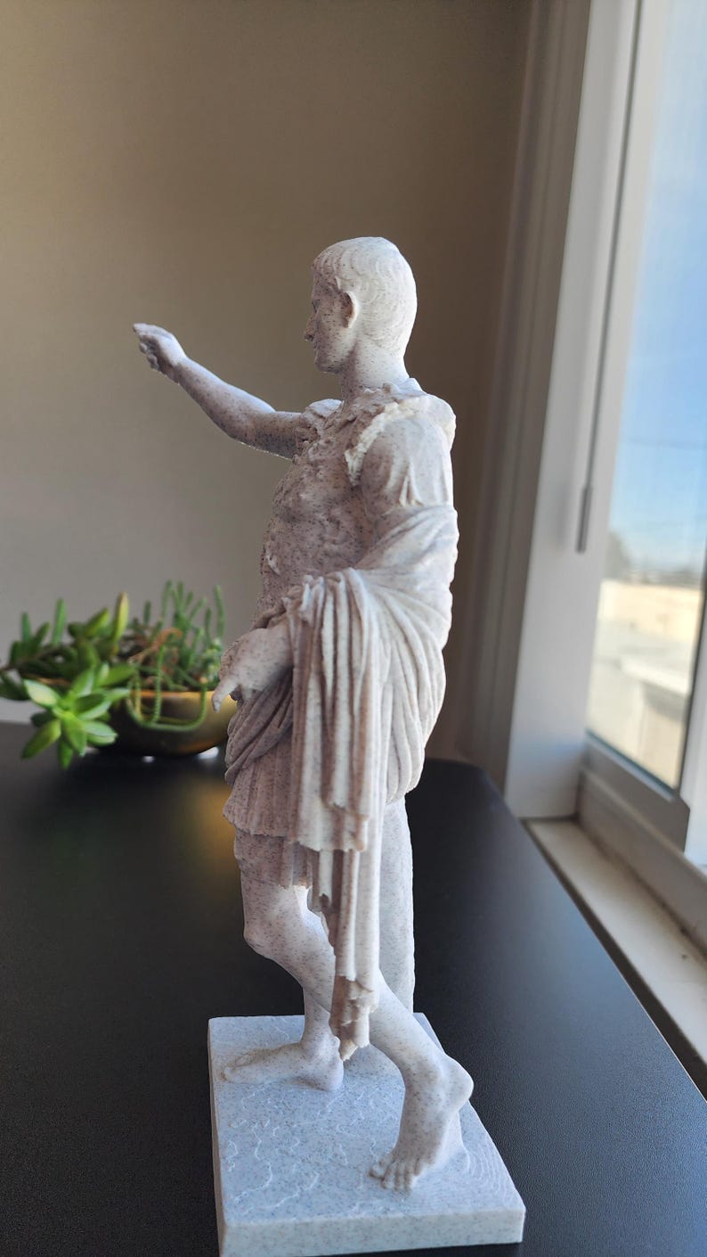 May include: A light-colored, speckled statue of a figure in Roman-style armor and a draped toga. The figure stands on a rectangular base with one arm extended forward and the other arm bent at the side, featuring detailed fabric folds and textures.