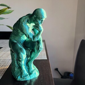 May include: A teal-colored sculpture of a seated figure, reminiscent of Rodin's "The Thinker." The statue is positioned on a dark surface, with a plant and a window in the background. The sculpture is approximately 10 inches tall.